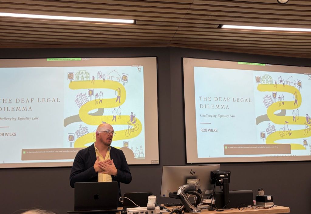 Rob Wilks speaking in British Sign Language at the launch of The Deaf Legal Dilemma, with his hands held to his chest and the book cover displayed on screens behind him.